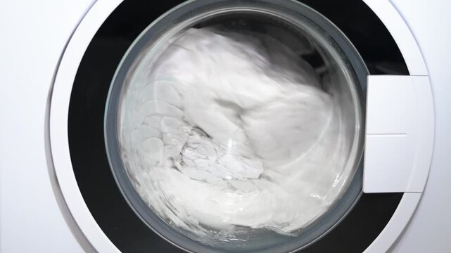 Washing machine cleaning and spinning white laundry. Close-up of a modern washing machine spinning and cleaning white clothes during a laundry cycle, with the drum rotating quickly with soapy water