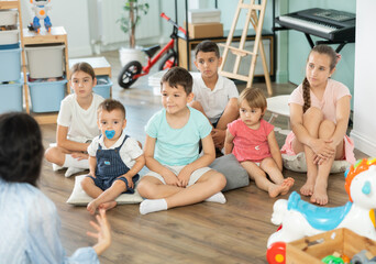 Obraz premium Boys and girls children sits on floor and listens to babysitter, organized pastime for children on summer holidays, kindergarten or day care activity