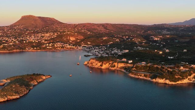 Aerial drone view of a flat-topped hill and a seaside village beside a bay at sunset, Chania, Greece