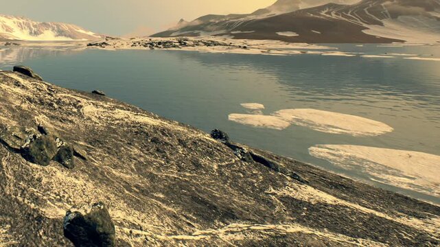 A large body of water reflecting nearby rocks and distant mountains during the early hours of the day. The surface features ice patches from melting snow.