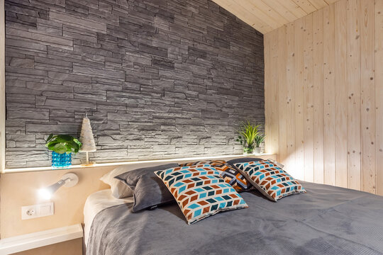 Cozy bedroom in a wooden house. The wall at the head of the bed is decorated with gray tiles under natural stone. The room is decorated with indoor plants.