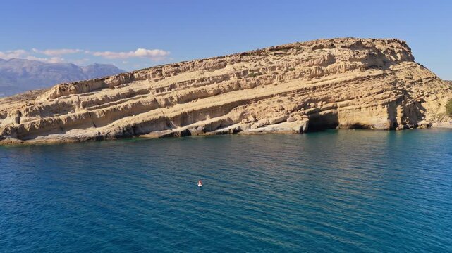 Aerial drone view of the layered sandstone cliff with weathered caves above the clear turquoise sea, Matala, Greece