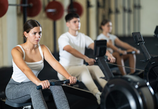 Workout girl cross training exercising cardio using rowing machine with group of people in crossfit club