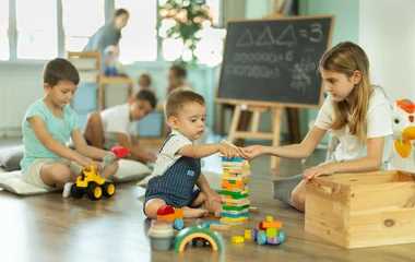 Boy and a girl of different ages sit together near a construction set and assemble it against the background of other children in kindergarten © JackF