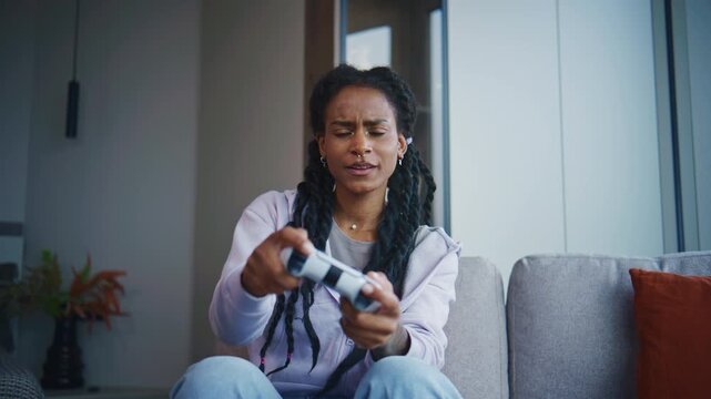 Woman sits on couch and plays video game with controller at home in modern living room during daytime