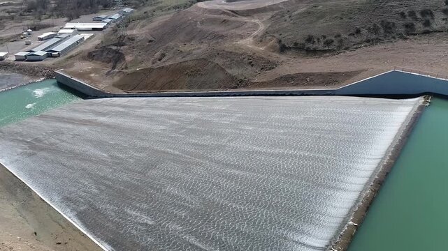 Aerial view of a dam spillway releasing water into the downstream channel beside a reservoir. Scenic hydraulic infrastructure surrounded by rugged mountains and clear turquoise water.