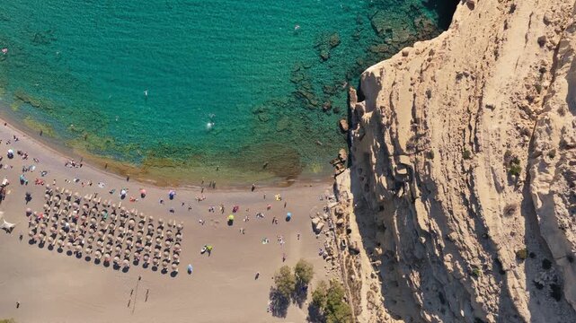 Overhead aerial drone view of Matala beach with straw umbrellas and clear turquoise water over a rocky seabed, Matala, Greece