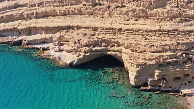 Aerial drone view of a pale sandstone cliff and wide sea cave above clear turquoise water, Matala, Greece