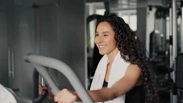 Smiling Hispanic Female Exercising On Elliptical Machine At Indoor Gym, Engaged And Motivated, Enjoying Workout, Maintaining Fitness And Health In A Positive Atmosphere.