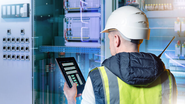 Industrial automation engineer monitors electrical control panels with tablet in smart factory control room ensuring diagnostics, maintenance and safety compliance