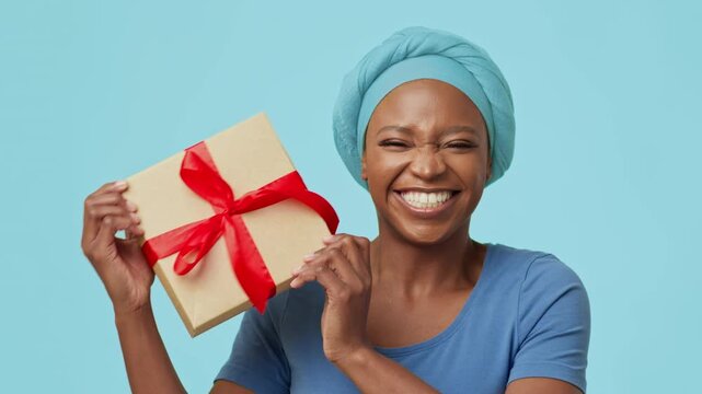 Smiling Black Woman In Blue Top Holding A Gift Box Tied With A Red Ribbon. Expressing Joy And Excitement While Celebrating Special Occasion In A Bright Studio Setting. Gift Concept.