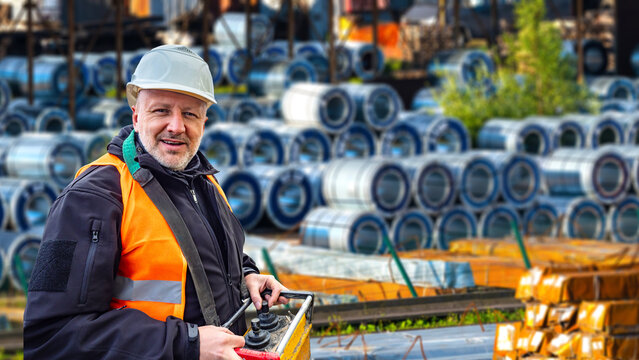 Steel coil storage yard operations worker controls crane handling system with remote controller in industrial metal warehouse facility