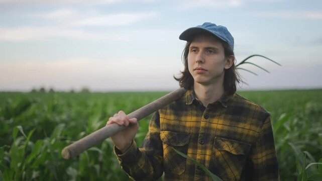 A young man stands in a field of corn, observing the crops. The corn sways gently in the breeze, while he holds a farming tool.
