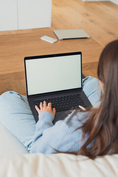A woman types on a laptop while sitting on a couch at home, wearing casual jeans and a cozy sweater. Blank screen offers space for mockups, remote work, study, browsing or creative projects in a relax