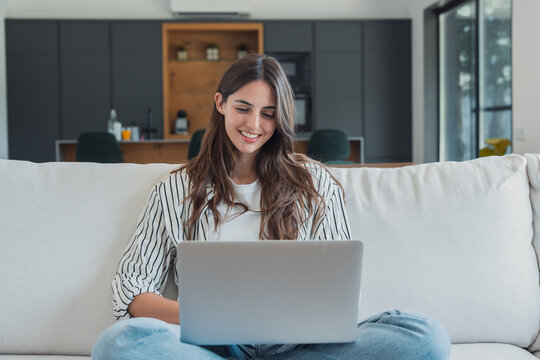 Young woman smiling, sitting cross-legged on a comfortable couch, using a laptop computer for remote work or online learning, enjoying the flexibility of a modern home environment