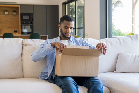 Young man sitting on a comfortable sofa in a modern apartment, opening a cardboard delivery box received from an online purchase, engaging in product unboxing
