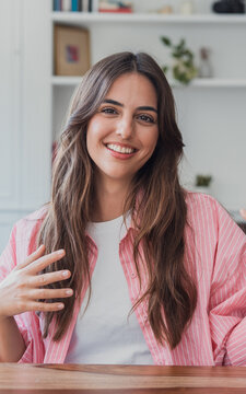 Smiling young woman with long brown hair sits at a desk in a bright home office, speaking and gesturing during a casual remote work video call. Warm, approachable, confident and natural portrait.