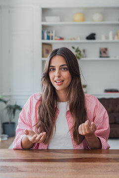Young woman speaks to camera while seated at a wooden desk in a bright living room, gesturing naturally during a casual video call or remote meeting, conveying confidence, warmth and approachability.