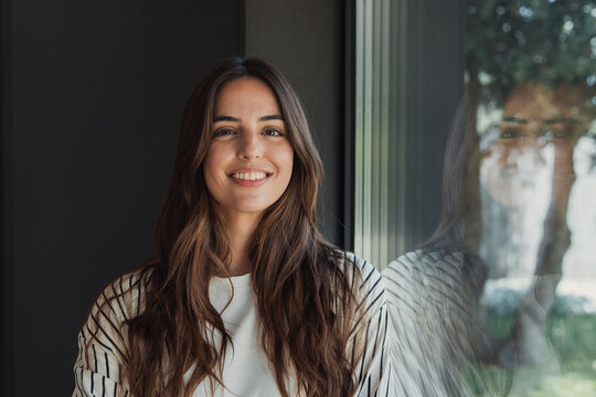 Young woman with long brown hair smiling at the camera, standing indoors next to a large window reflecting trees and nature, wearing a striped shirt