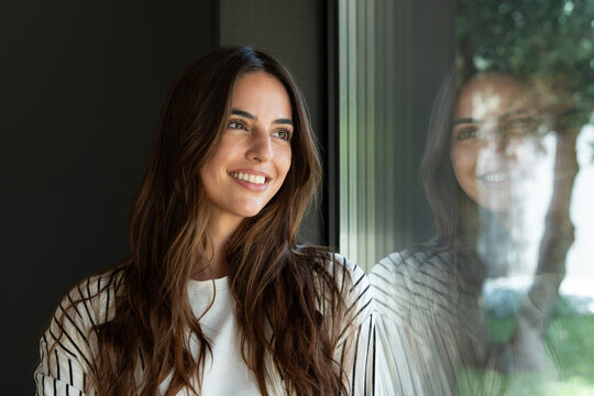 Young woman smiling with genuine happiness, looking away through a reflective glass window inside an indoor space, showing her reflection
