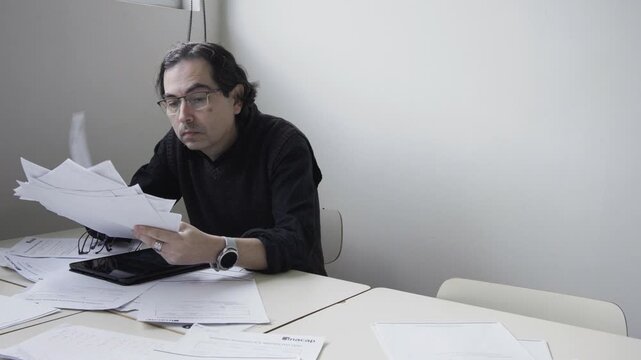 Tired male teacher taking off glasses, rubbing his eyes, and massaging his neck in deep exhaustion at a desk