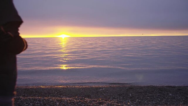 Rear view of a man walking onto a rocky shore to watch a beautiful golden sunrise over the ocean