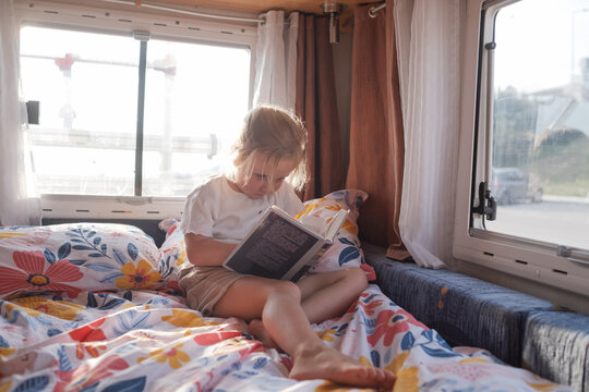  Focused child reading a storybook on a motorhome bed during a sunny summer family road trip.