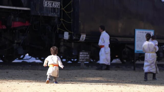 Child walks toward historic train with curious wonder. Adults in white robes stand nearby, observing. Train rests quietly, its metal gleaming. Sunlight falls across dusty ground