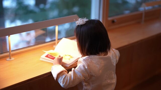 Soft light glows on a child's reading page. Little girl sits quietly, engrossed in her book. Hair adorned with a delicate flower. Warm glow illuminates her focused expression. Ideal for quiet learning