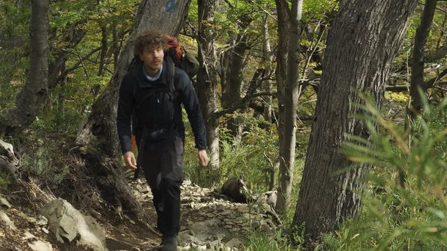 Hiker with a backpack walking along a dirt trail in a forest, surrounded by trees with autumn leaves, outdoor activity.