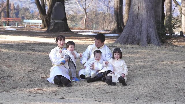 Family sits under large tree in park. Children wear white traditional outfits. Parents smile, gently engaging with kids. Scene feels peaceful and warm. Ideal for family, cultural, or seasonal themes