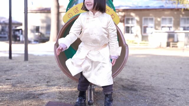 Child sits inside green tunnel playground structure. Brightly dressed toddler smiles, holding on with both hands. Sunlit park with sandy ground, distant buildings, and bare trees