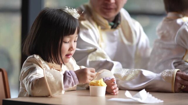 Little girl eats dessert with eyes closed. Baby sits beside her, eyes wide open. Adults watch warmly from behind. Gentle family moment in cozy, softly lit room