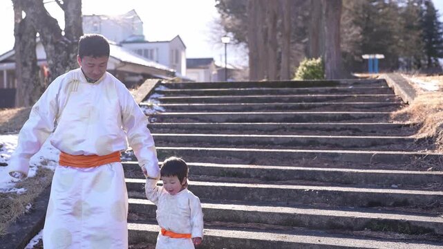 Man and child ascend stone stairs in traditional robes. Winter sun illuminates snowy patches on steps. Child holds man's hand, showing trust and comfort. Scene evokes quiet tradition and familial bond