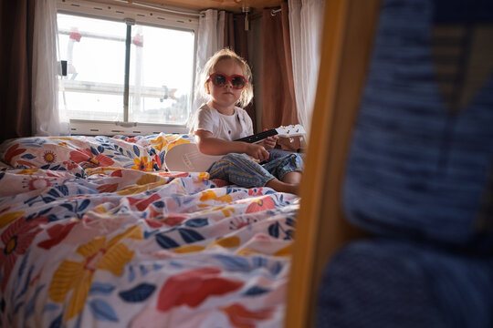 Cool little girl sitting cross-legged on a bed and playing a white ukulele in a sunlit campervan.