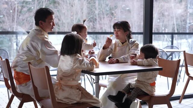 Family gathers at table in bright, modern cafe. Children dressed in matching traditional attire. Parents smile, children laugh, enjoying shared meal. Natural light streams through large windows