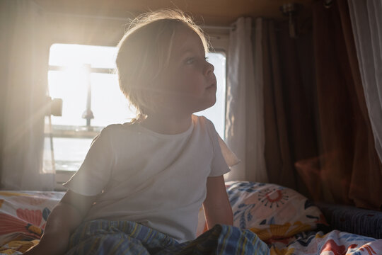  little blonde girl lying on a cozy bed inside a campervan and looking out the bright sunlit window.