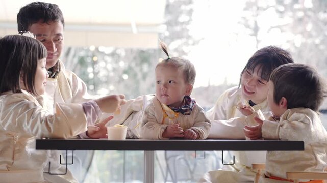 Family shares joyful meal at cozy table. Children laugh, reach, and feed each other. Warm lighting highlights tender, happy moments. Perfect for family, lifestyle, or cultural content