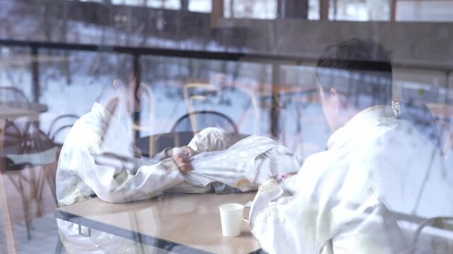 Warm light spills through a winter cafe window. Man in white sits, hands clasped around a cup. His reflection dances on the glass, soft and blurred. Inside, snow blankets the outside streets