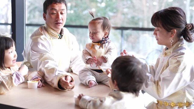 Joyful family gathers around a table. Children and a baby share smiles and laughter. Parents guide the moment with affection. Bright daylight streams through large windows. Ideal for family, parenting