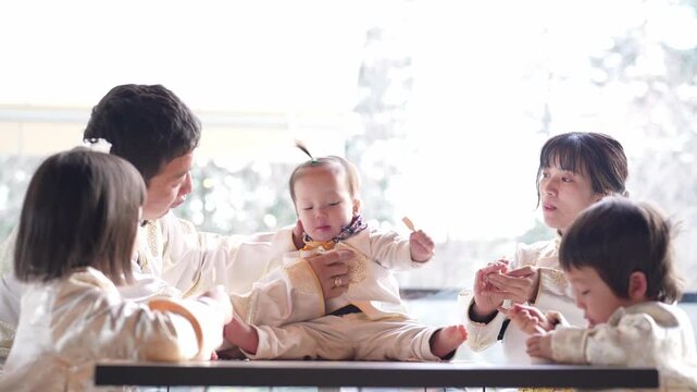 Warm sunlight bathes a family gathered around a table. Parents and three children wear matching outfits. Baby sits in the center, surrounded by adoring faces. Everyone smiles, sharing a tender