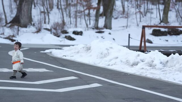 Little child walks snowy road alone. White coat contrasts with gray asphalt and snow. Child moves calmly across painted crosswalk. Winter forest looms softly in background. Calm, quiet moment in cold
