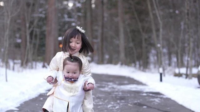 Snowy path winds through tall trees. Two children, dressed in white, walk side-by-side. Third child follows behind, wearing a white robe. They smile, enjoying the winter day