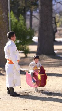 Sunlight glints off a man's white robe. Child laughs, clinging to a red rocking horse. The man steadies the toy, smiling gently. Trees loom behind, casting soft shadows