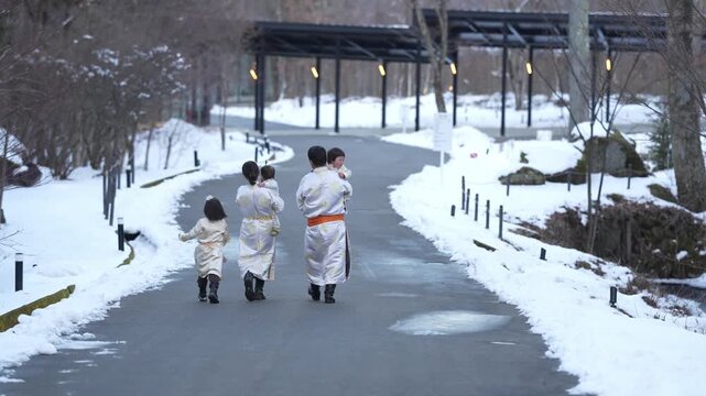 Snow blankets a quiet garden path. Four people in traditional robes walk forward. They move with calm purpose, heads slightly bowed. Warm lanterns glow softly under a covered walkway