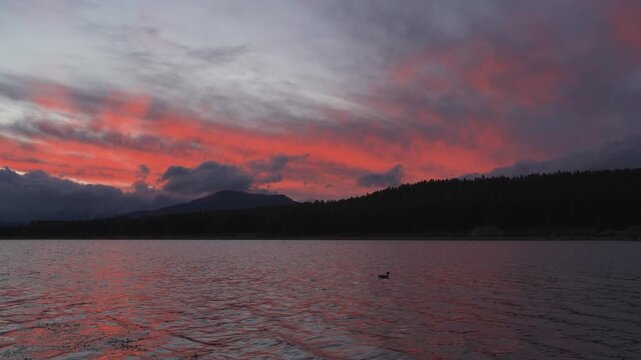 Dramatic sunset with vibrant red and purple clouds reflecting over the calm water and silhouetted mountains of Laguna La Zeta, Esquel, Chubut, Argentina.