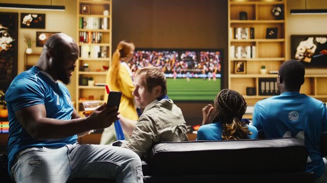 A man on an apartment couch holds a smartphone and tells friends about negative online match results while soccer supporters drink beer at home.