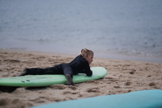 Little blonde girl in a black wetsuit lying on her stomach on a green surfboard on a sandy beach,