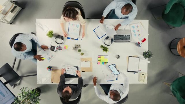 Office team is shown from a close overhead angle in multinational office, planning resources and budgets around performance metrics, data review, report sharing and planning session.