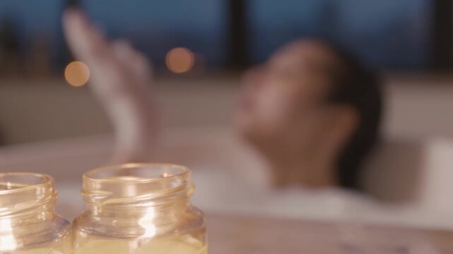 Aromatic candles lit with a blurred background of a young woman in the bathtub. Medium shot capturing the tranquil spa corner during sunset focusing on aromatic candles by the hot tubs edge.